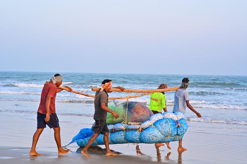 Four fishermen are carrying their gear along the beach towards the sea.