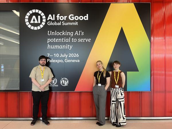Three adults stand in front of a large banner that reads AI for Good with a large multicolour stylised letter A against a black background.